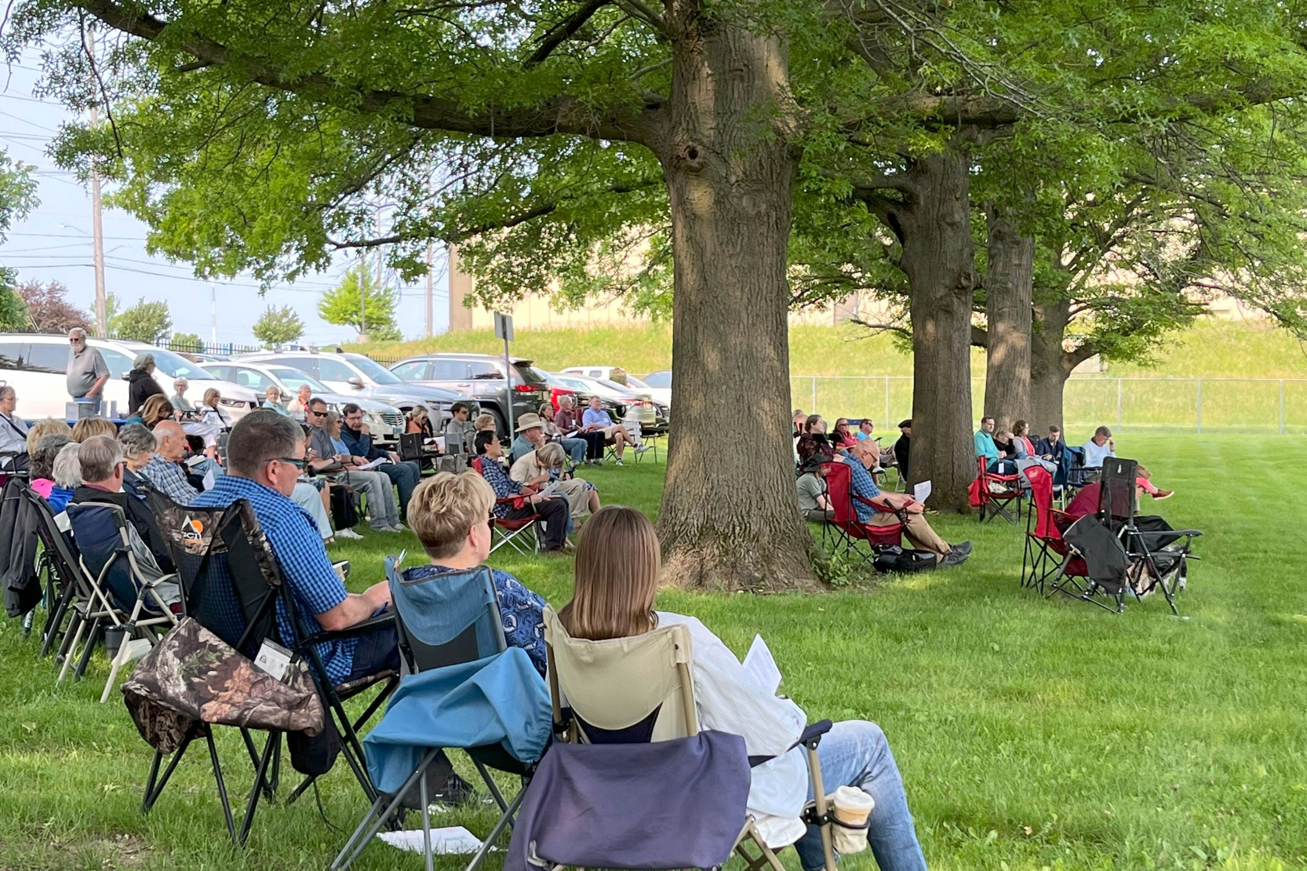 View of worship participants for Summer 2025 Outdoor Sunday service. People sitting in semi-circle of lawn chairs placed on grass under large trees.