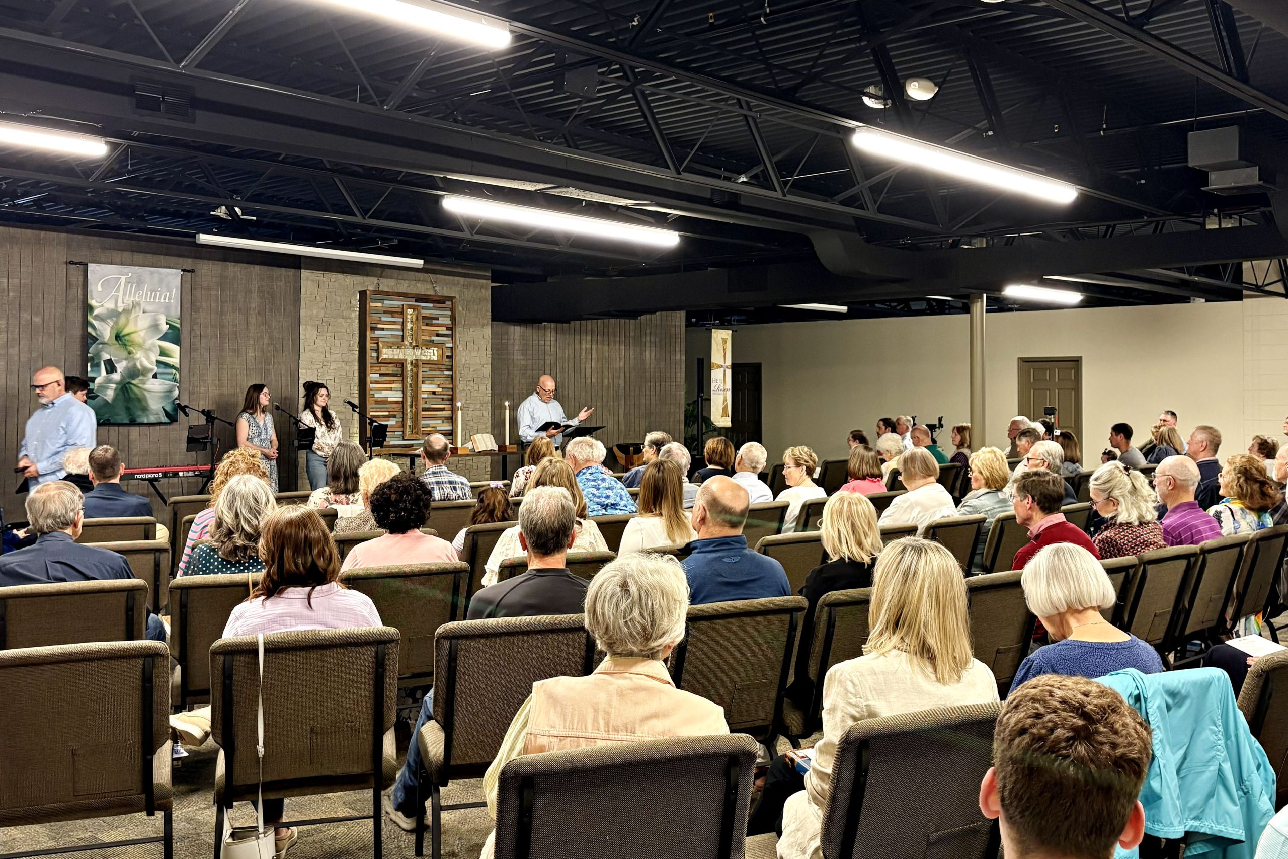 2026 first worship 0412lo first worship in new location for Faith Lutheran Church of McLean County (Faith Church) in Bloomington Illinois. Image is many rows of people sitting in chairs facing a cross listening to the preacher