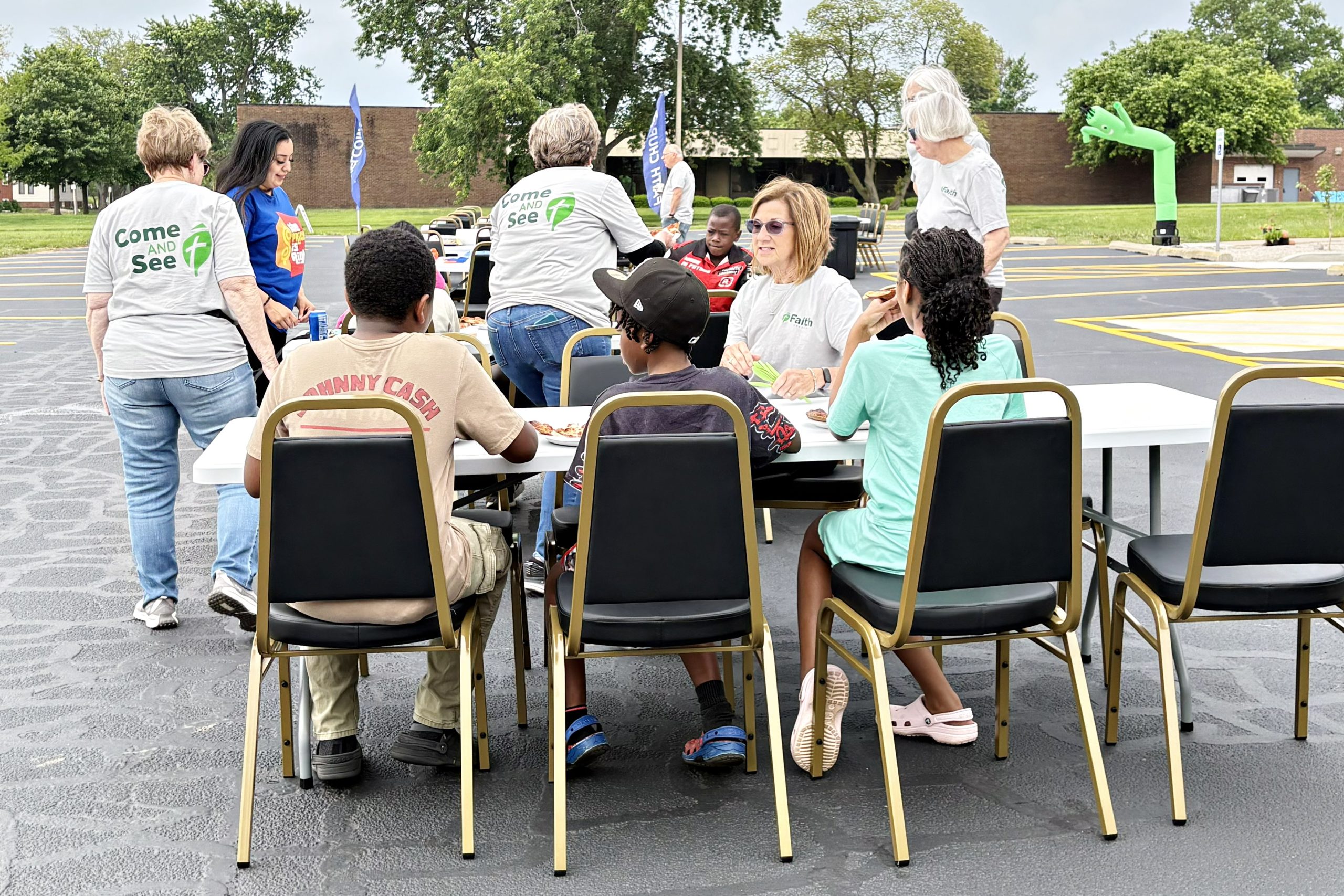 2026 0516 FT b neighbors enjoying an outdoor lunch with one another seated at a rectangualr table in Faith Lutheran Churc of McLean County's Bloomington parking lot